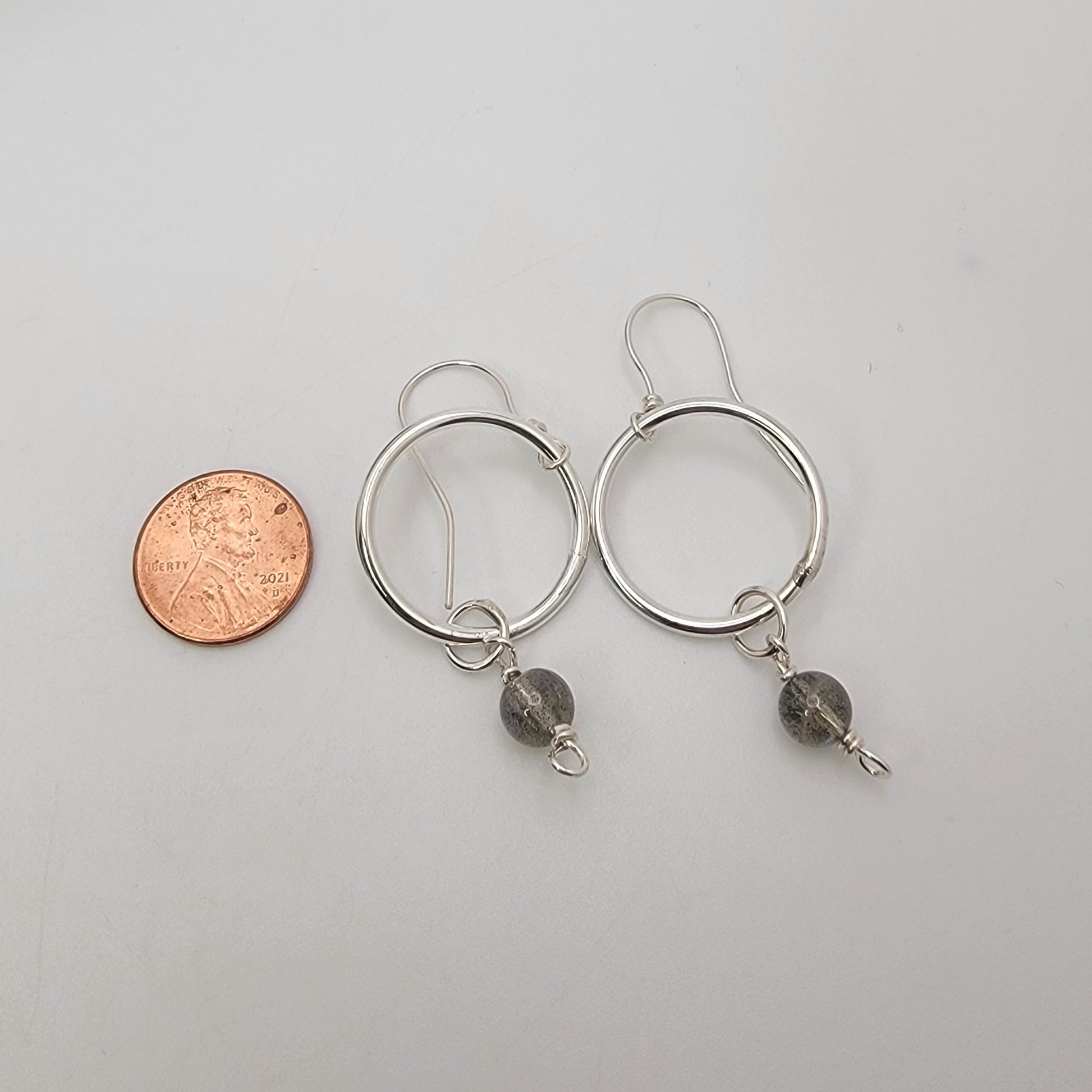 A pair of silver-toned hoop earrings with small dark gray bead drops, shown next to a penny for scale on a light gray background.