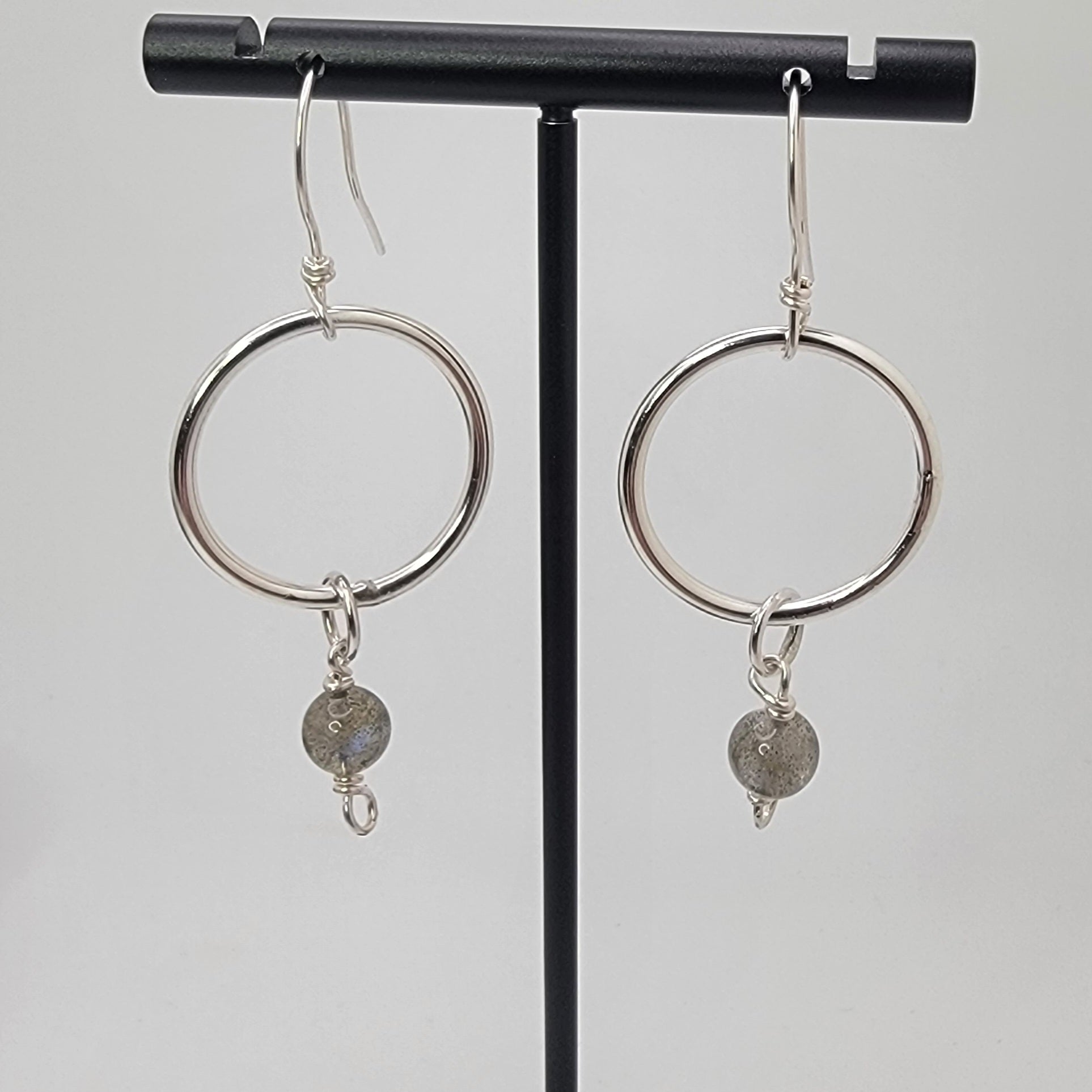A pair of silver hoop dangle earrings with small gray stone charms, displayed on a black jewelry stand against a light gray background.