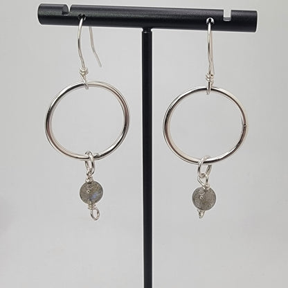 A pair of silver hoop dangle earrings with small gray stone charms, displayed on a black jewelry stand against a light gray background.