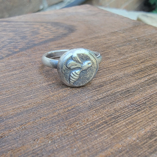 A silver-toned ring featuring a circular medallion with an embossed bee design, resting on a rustic wooden surface.