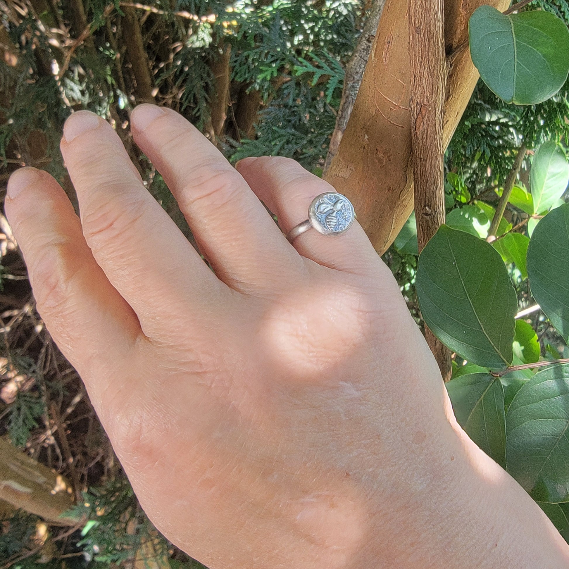 A silver ring with a round, textured bezel-set stone worn on a person's ring finger, shown against a background of green foliage and tree trunks.
