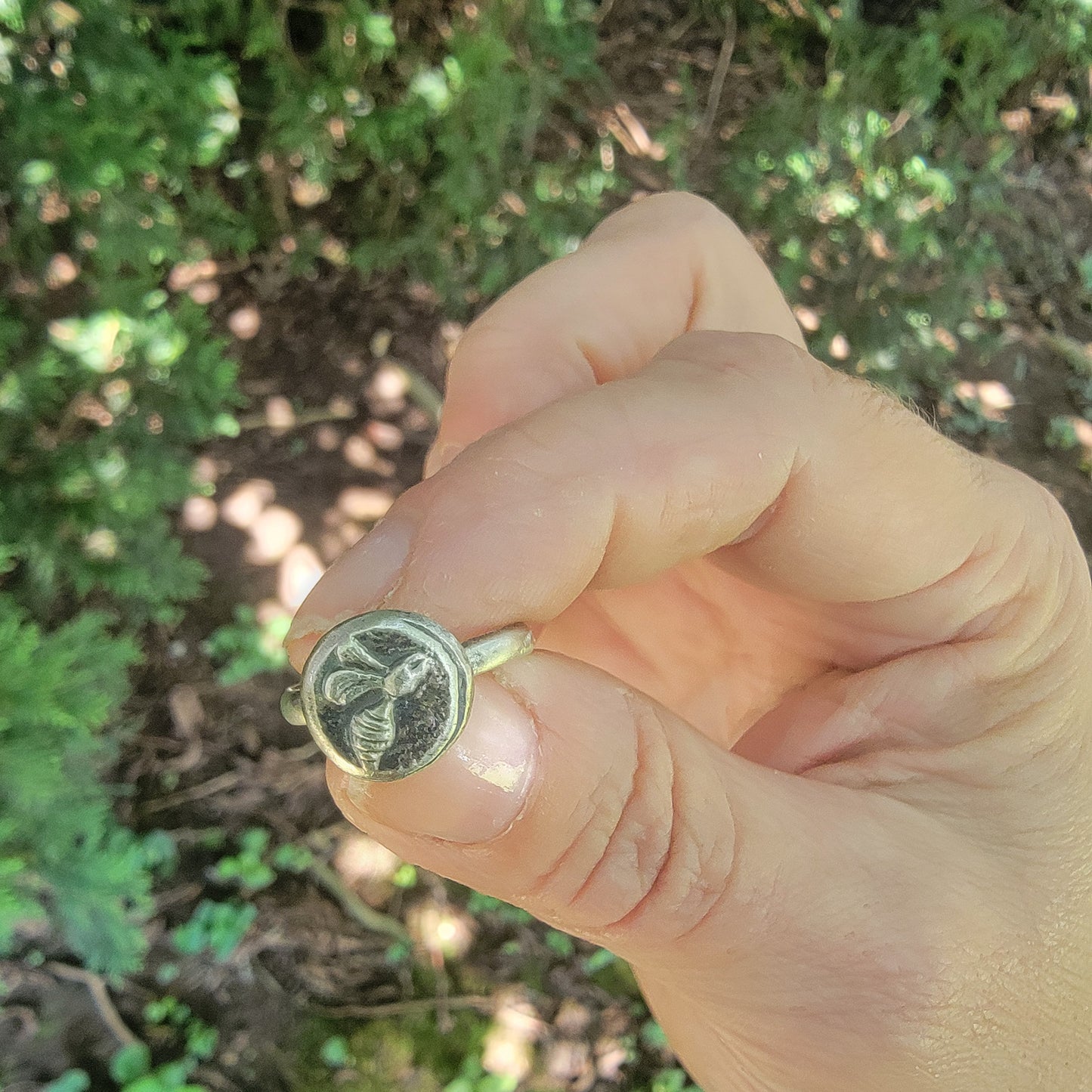 A hand holding a silver signet ring with an engraved bee design, set against a blurred outdoor background of green foliage.