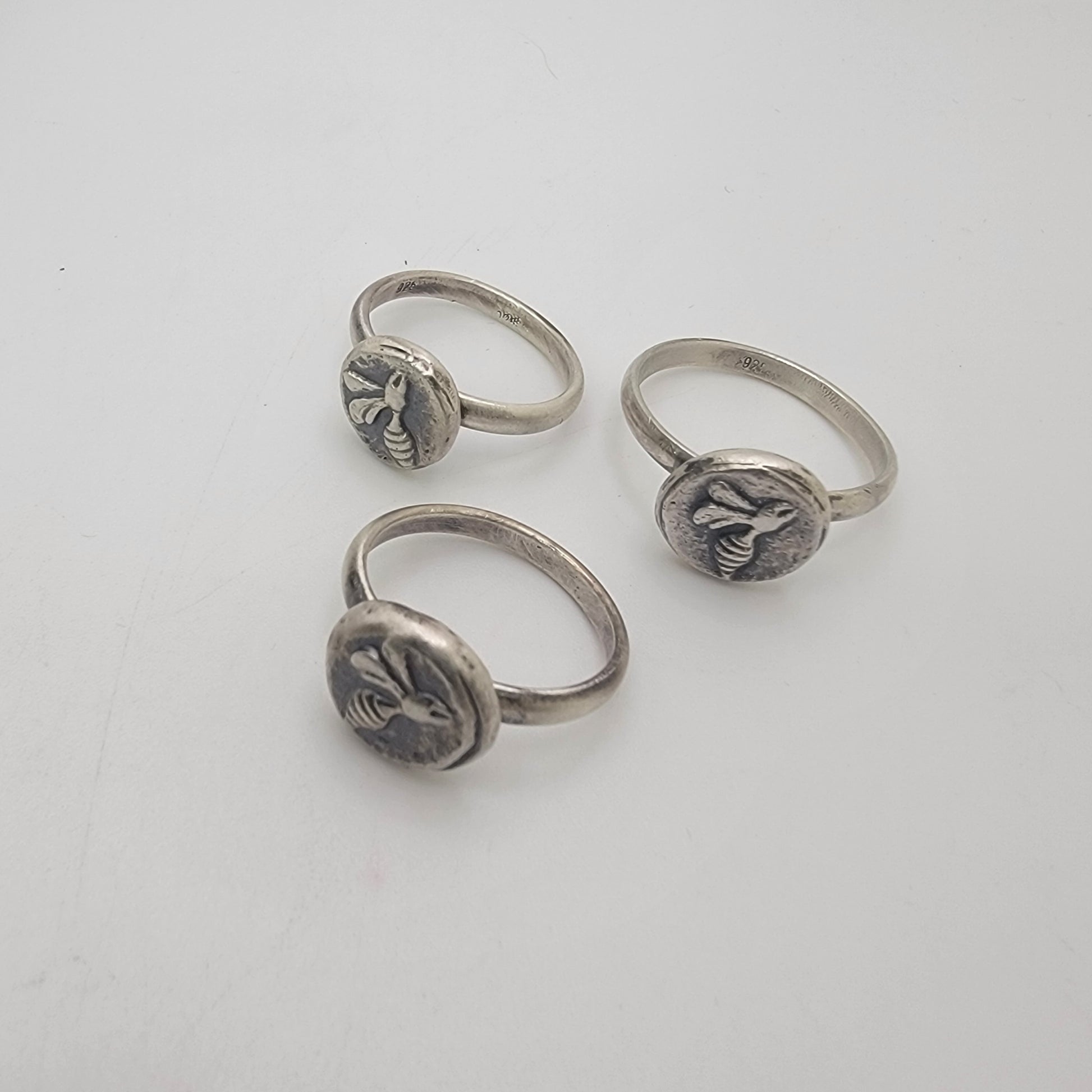 Three silver rings with round bezel-set bee motifs, displayed on a light gray background.