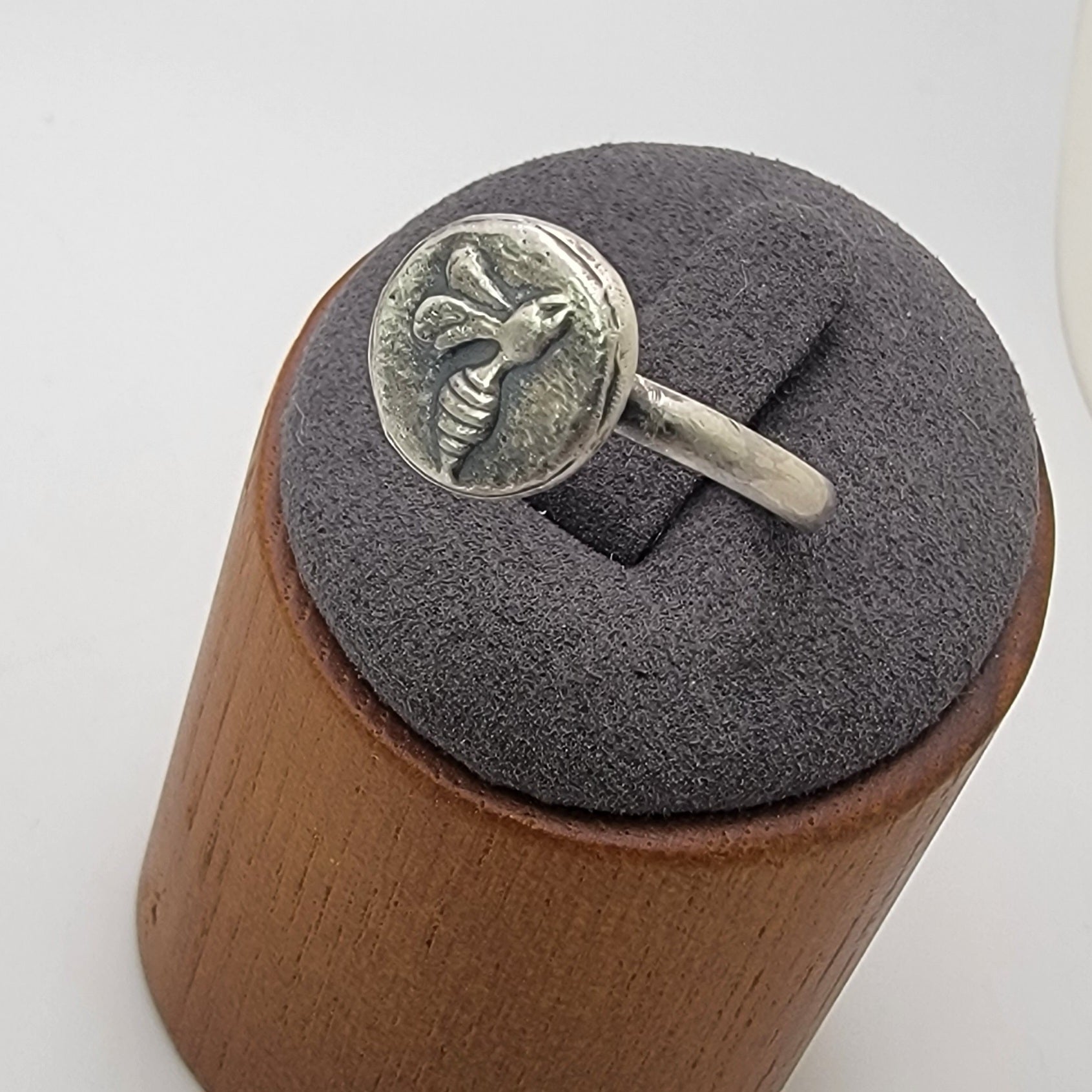 A silver ring with an embossed bee motif on a circular face, displayed on a gray velvet cushion atop a wooden stand against a white background.