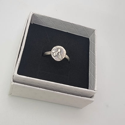 A silver ring with an engraved bee design on a round face, presented inside an open light gray jewelry box with a black velvet insert.