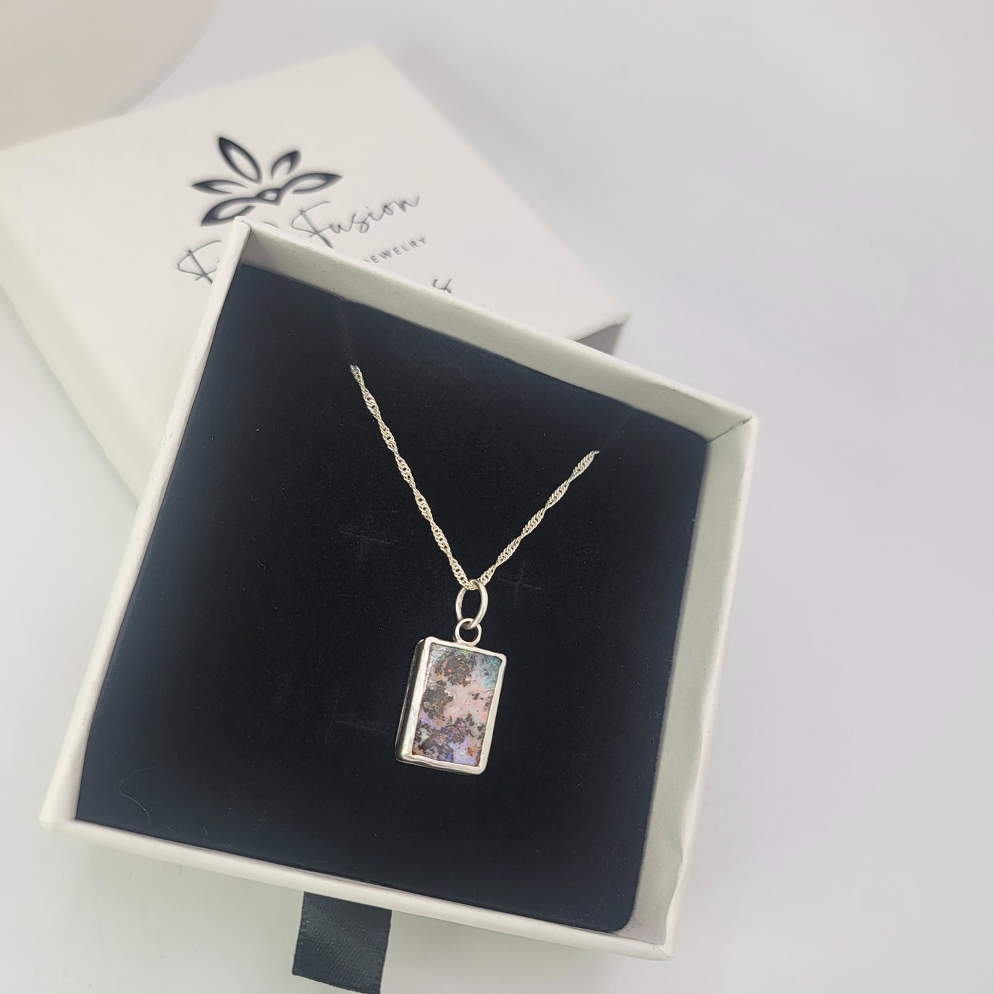 A silver twisted chain necklace with a rectangular boulder opal pendant, presented in an open white jewelry box with a black velvet insert. The brand box lid is visible in the background.