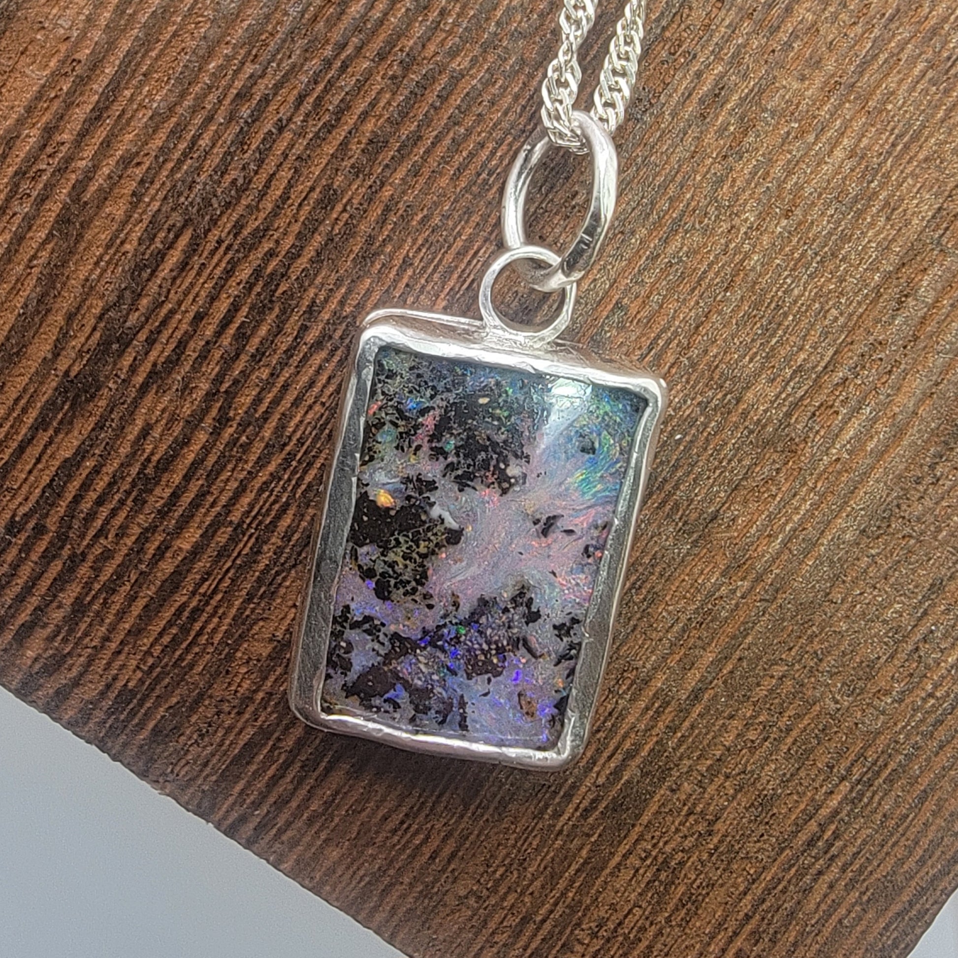 A silver rectangular pendant necklace featuring an iridescent opal stone with black and multicolor patterns, resting on a textured brown wooden surface.