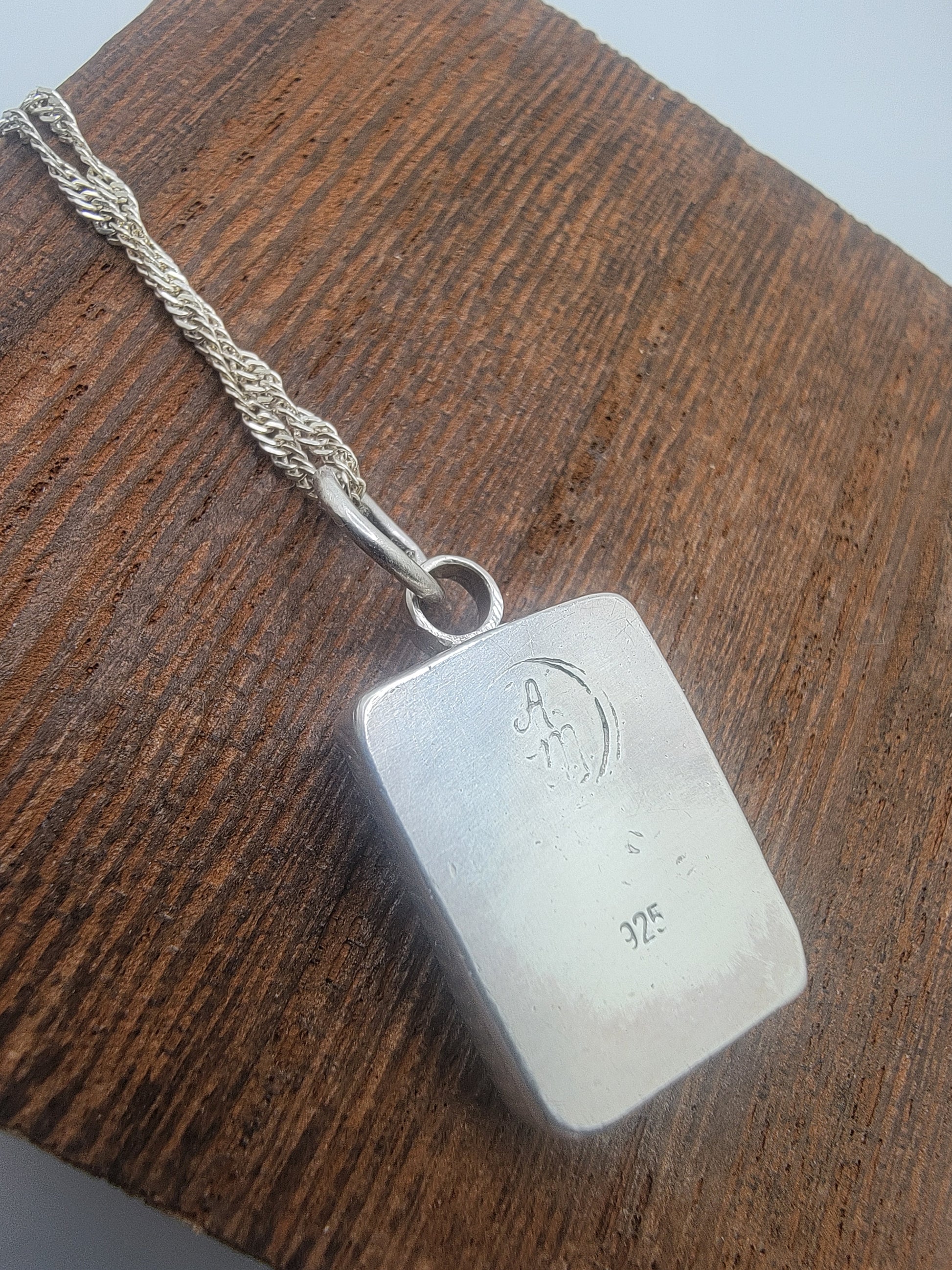 A sterling silver pendant necklace with a rectangular silver pendant engraved with maker's initials and '325', resting on a dark wood surface.