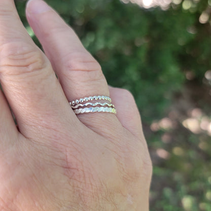 A close-up of a hand wearing three thin silver wavy bands rings, set against a blurred green outdoor background.