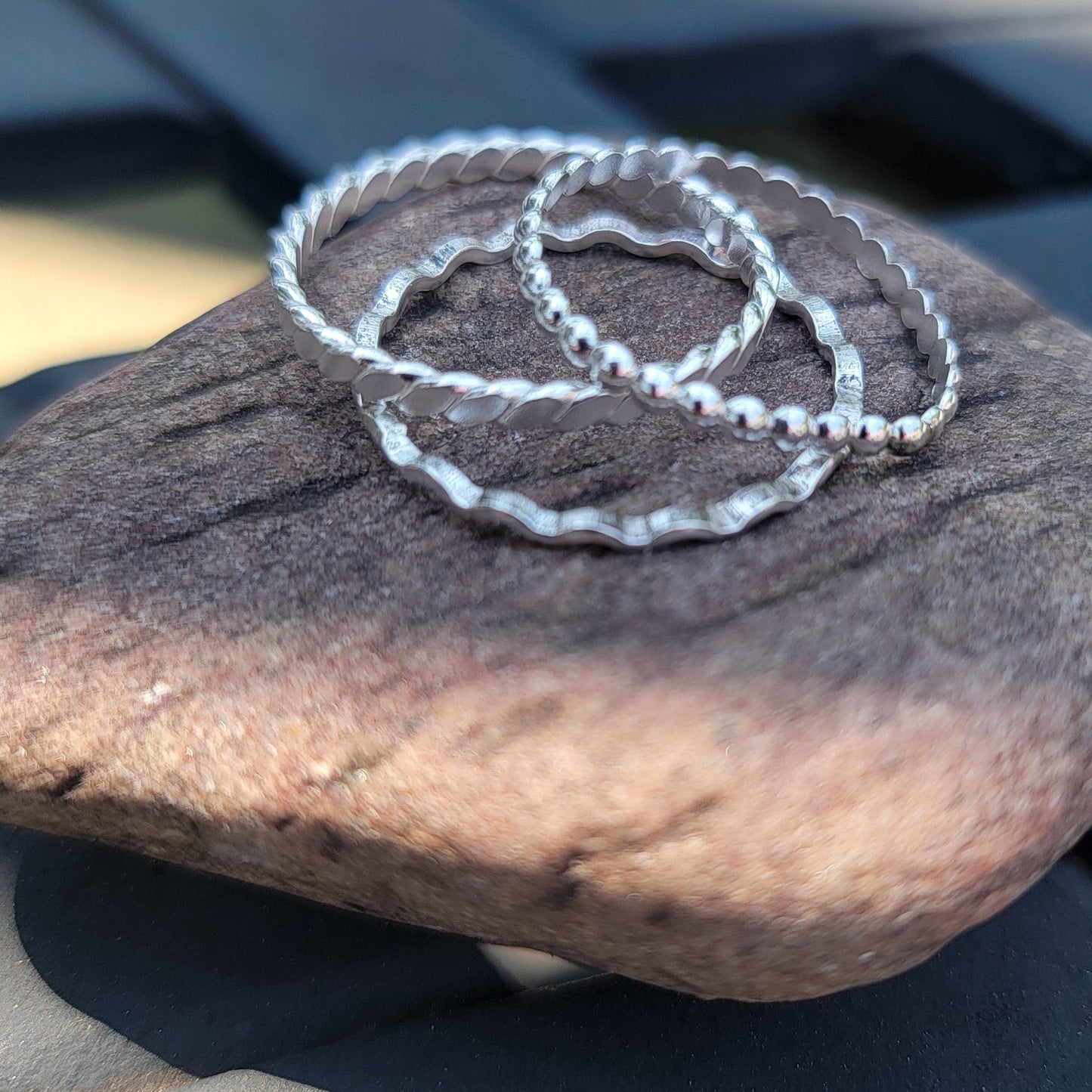 Three thin silver rings with a textured, hammered finish resting on a smooth brown stone, placed on a dark gray slatted outdoor table.