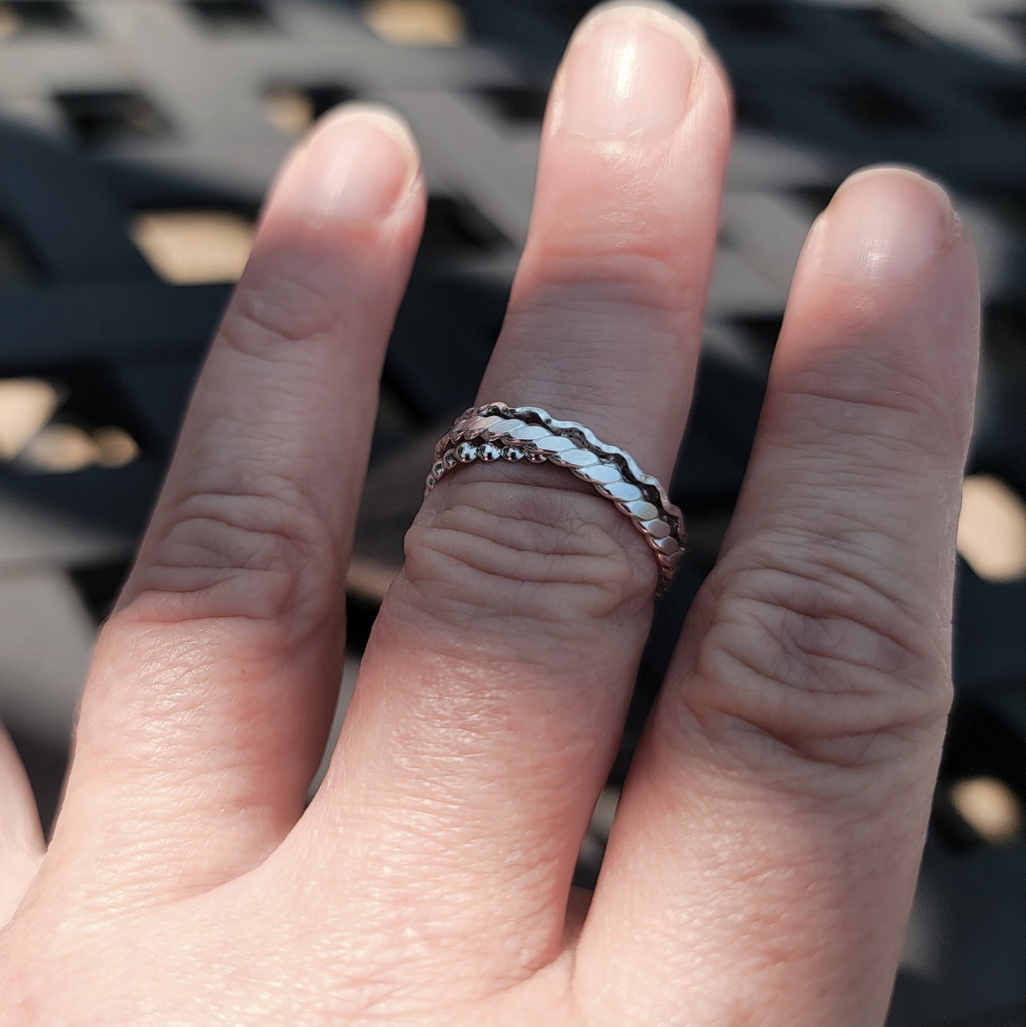 A close-up of a hand wearing three thin silver rings with wavy, textured patterns, set against a blurred black metal lattice background.