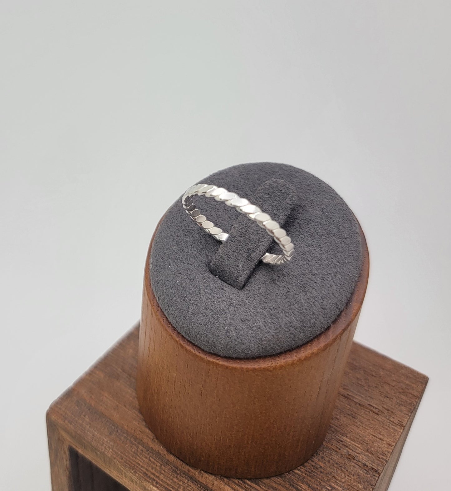 A silver twisted rope ring displayed on a gray velvet ring stand, which sits on a wooden block against a light gray background.