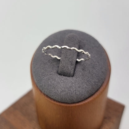 A delicate silver ring with a wavy, scalloped edge design, displayed on a gray velvet ring stand set inside a wooden cylindrical holder against a light gray background.