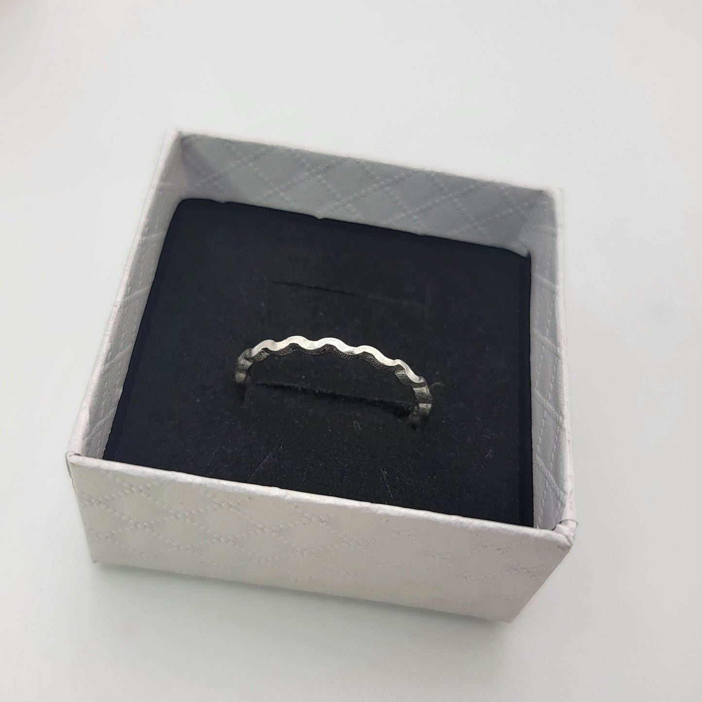 A silver ring with a wavy, scalloped edge displayed on a black velvet insert inside a square white gift box.