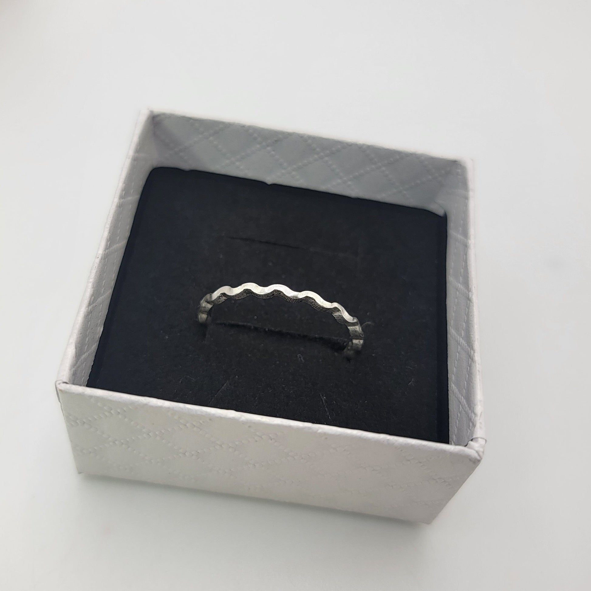 A silver ring with a wavy, scalloped edge displayed on a black velvet insert inside a square white gift box.