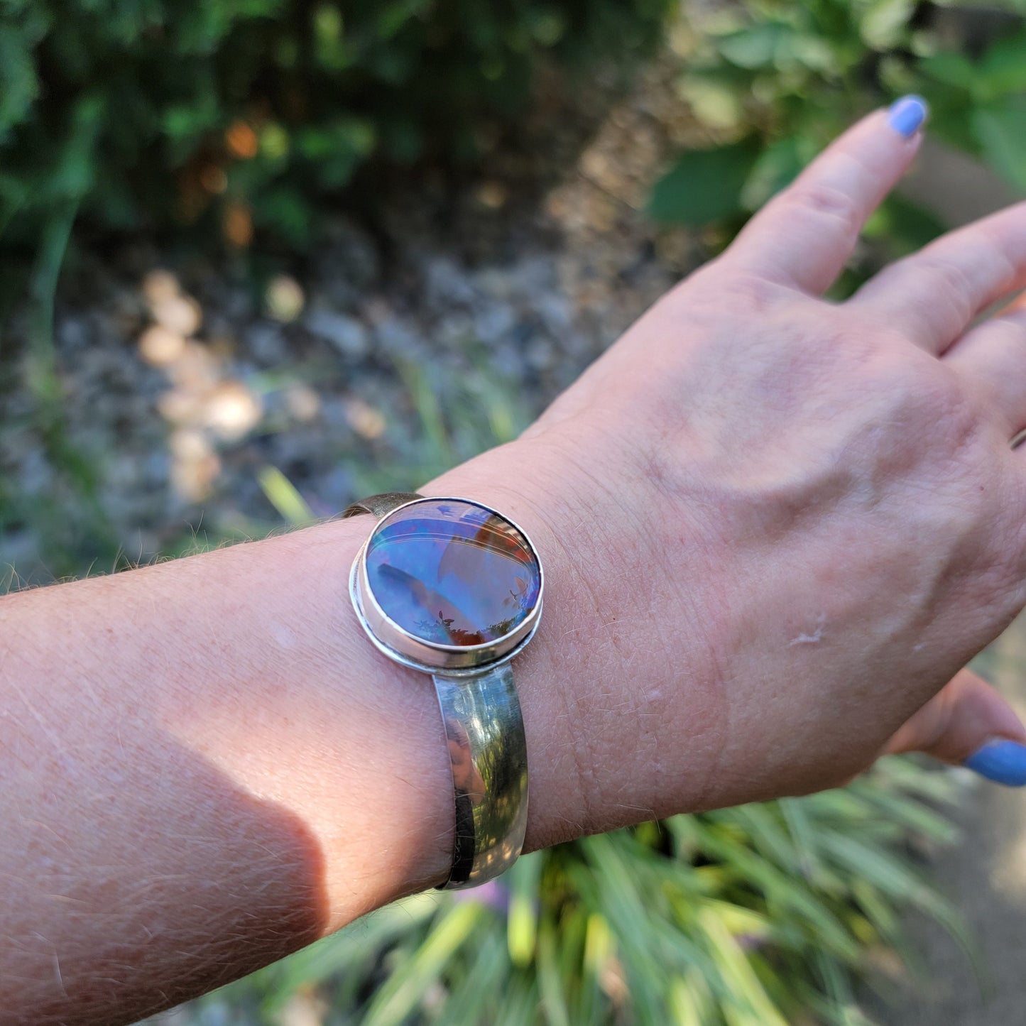 A silver cuff bracelet with a large round cabochon gemstone featuring blue, purple, and orange hues, shown worn on a person's wrist against a blurred garden background.