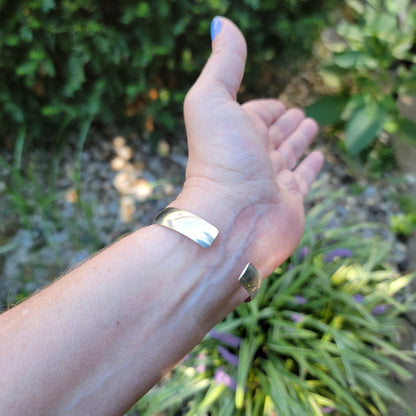 A person's hand and forearm wearing a wide, polished silver-toned metal cuff bracelet in an outdoor garden setting.