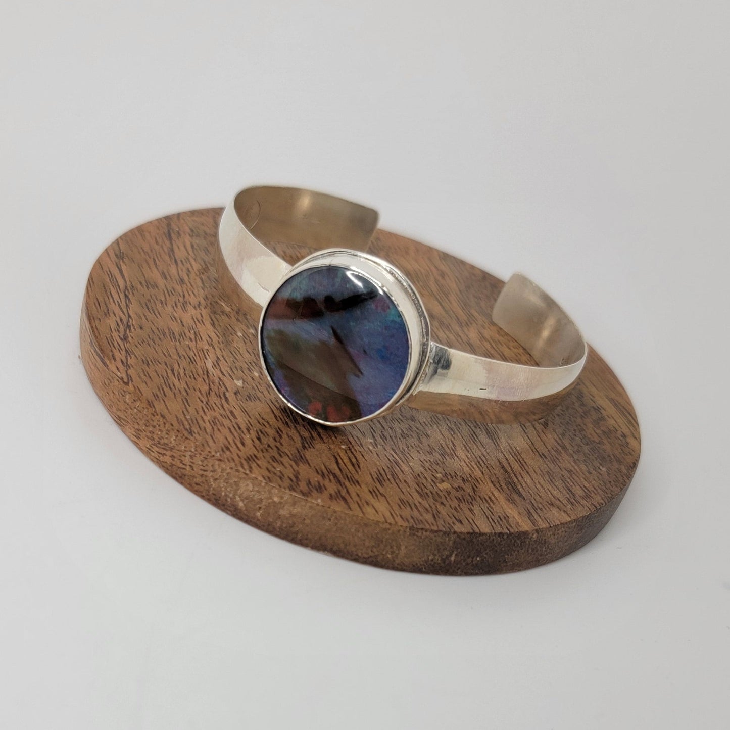 A silver-toned open cuff ring with a round dark blue and black stone, displayed on a small round wooden stand against a plain light background.