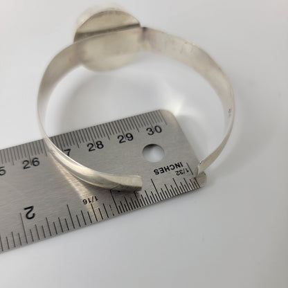 A silver-toned metal cuff bracelet with a circular top detail, shown next to a metal ruler for scale on a plain white background.