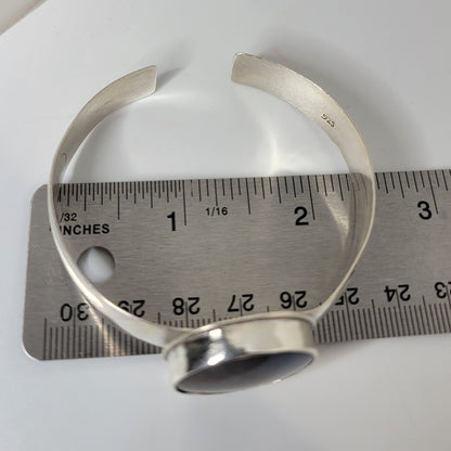 A silver-toned open cuff bracelet with a polished dome stone, shown next to a metal ruler for scale on a white surface.