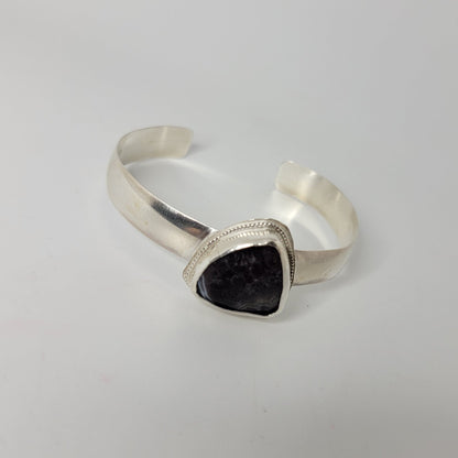 A silver cuff bracelet with a triangular bezel-set dark purple stone, photographed on a plain white surface.