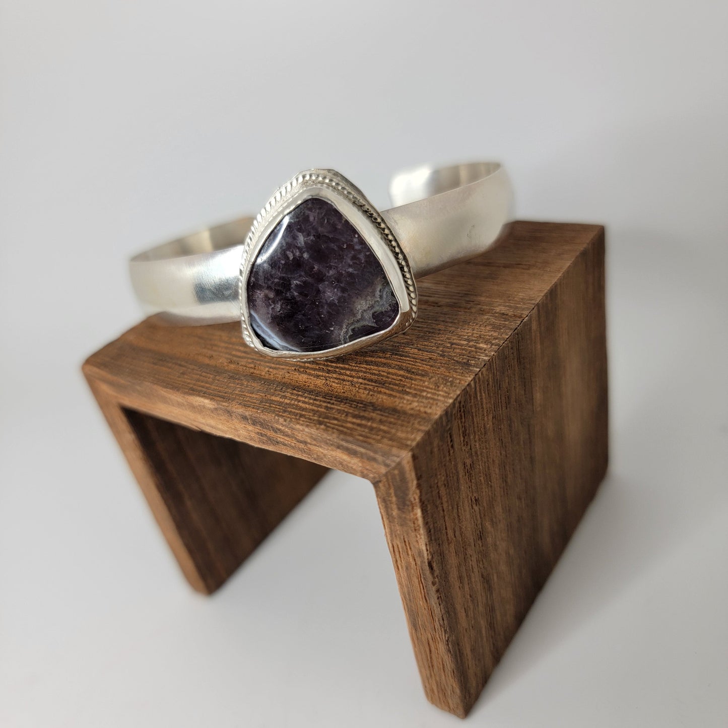 A silver cuff bracelet with a large triangle-shaped dark purple stone, displayed on a small wooden block against a light gray background.