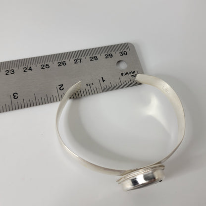 A silver cuff bracelet, shown next to a metal ruler for scale on a plain white background.