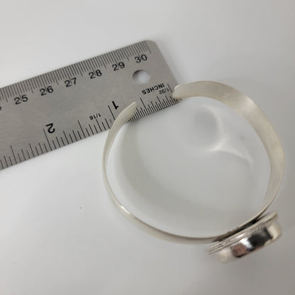A silver cuff bracelet with bezel set cabochon shown on a white background, with a metal ruler placed beside it for scale.