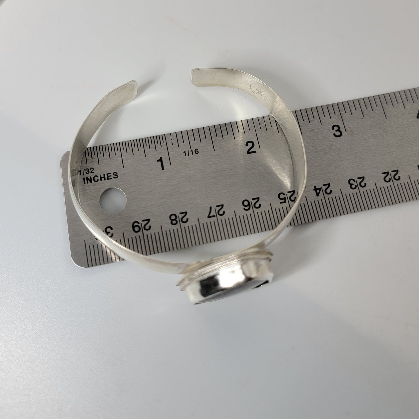 A silver open cuff bracelet is shown next to a metal ruler for scale on a white surface.