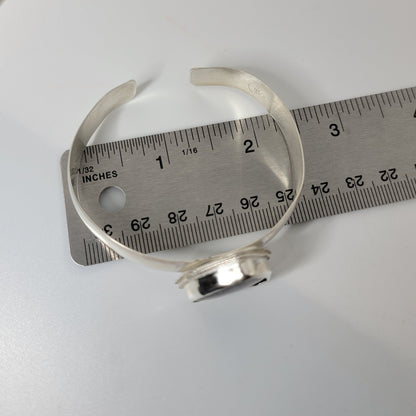 A silver open cuff bracelet is shown next to a metal ruler for scale on a white surface.
