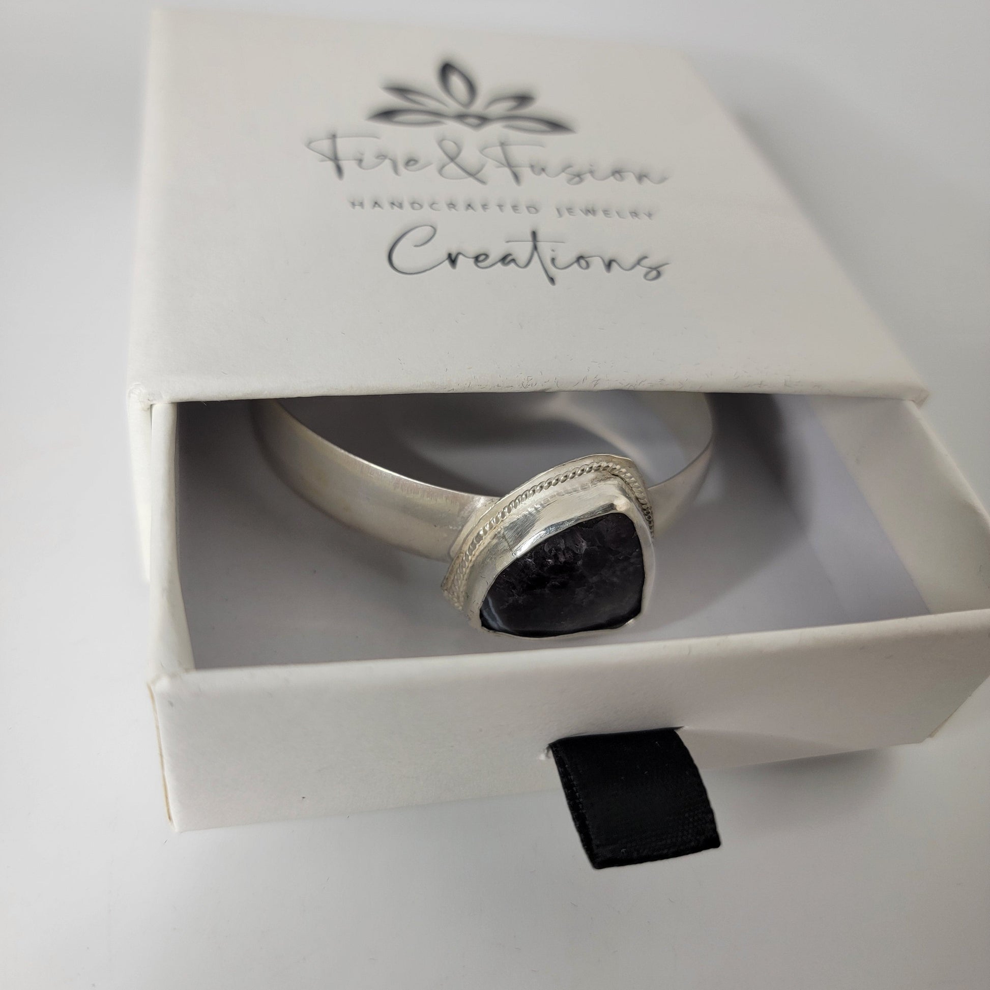 A silver cuff bracelet with a dark, textured square stone set in a silver bezel, presented inside a white gift box with the brand name 'Fire & Fusion Handcrafted Jewelry Creations' printed on the lid.