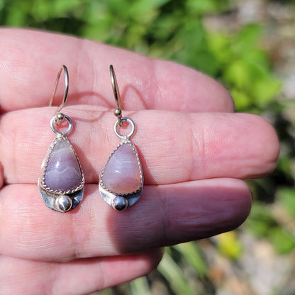 A pair of handmade silver dangle earrings with lavender-purple stone pendants, held in a person's hand against a blurred green outdoor background.