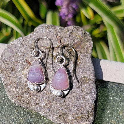 A pair of handmade dangle earrings with teardrop-shaped pinkish-purple stone pendants and silver hooks, displayed on a textured gray stone with green foliage in the background.