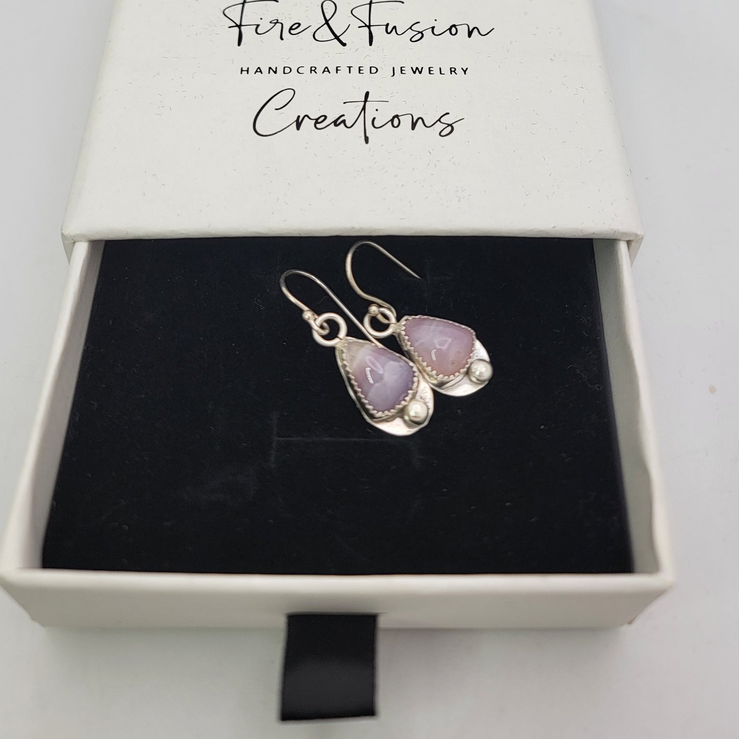 A pair of silver drop earrings with lavender purple teardrop gemstones and small pearl accents, presented in a black velvet-lined white jewelry box by Fire & Fusion Handcrafted Jewelry Creations.