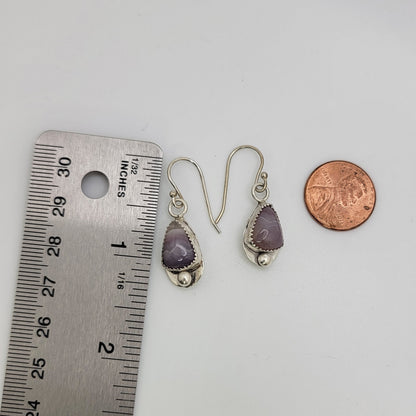 A pair of silver drop earrings with teardrop-shaped purple amethyst stones, shown next to a metal ruler for scale and a copper penny for size reference on a plain white background.