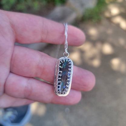 A hand holding a silver pendant necklace with a rectangular iridescent stone set in a textured silver border, with a blurred outdoor background.