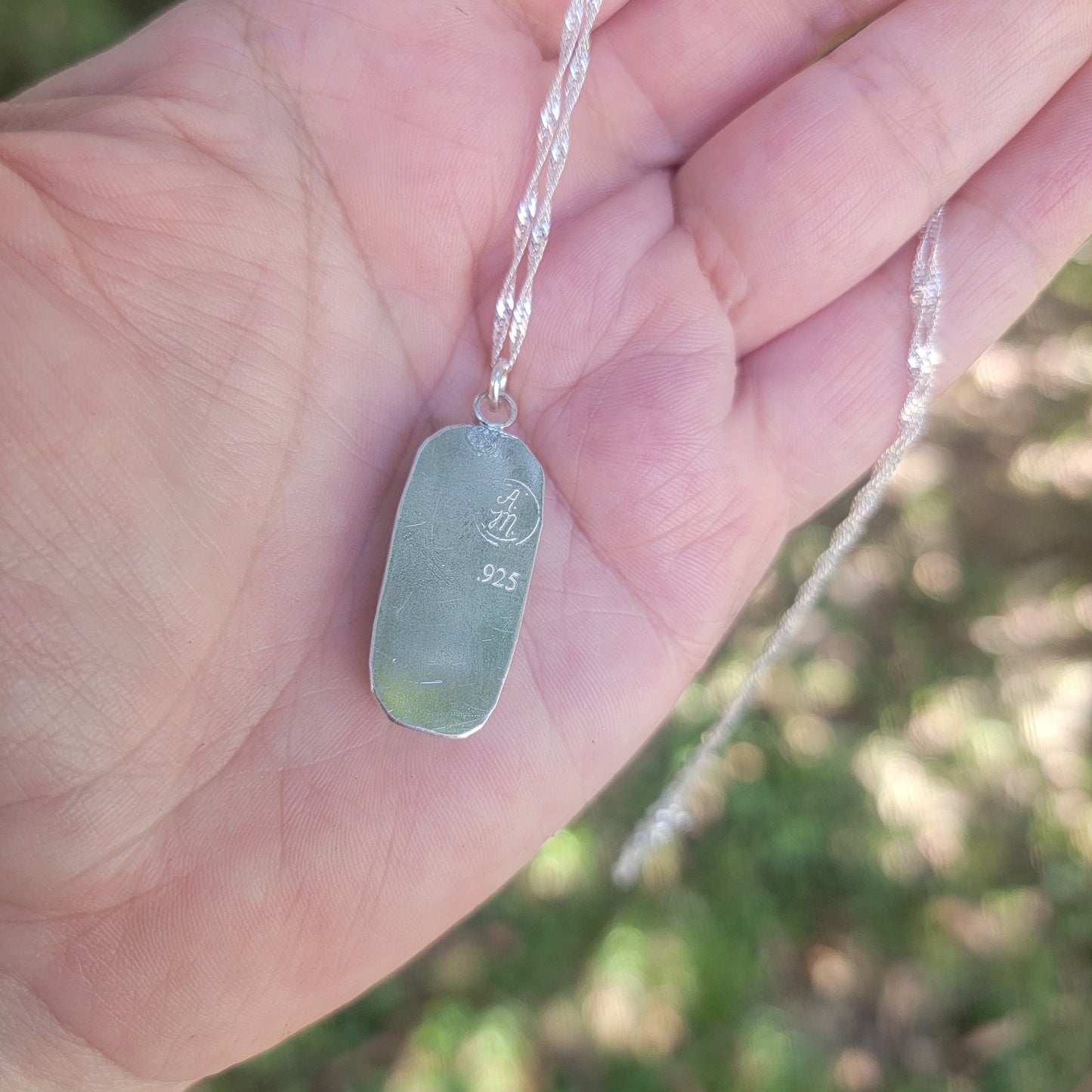 A sterling silver dog tag pendant engraved with makers stamp and '925' silver purity stamp, resting on a person's palm with a blurred outdoor background.