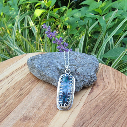 A silver-toned rectangular pendant necklace with a blue and green iridescent stone, displayed on a gray rock on a wooden surface with green foliage in the background.