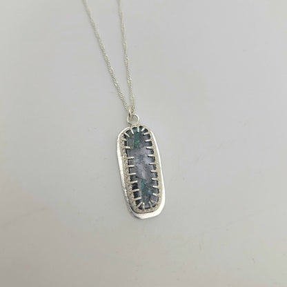 A silver pendant necklace featuring a rectangular, openwork silver frame holding a central oval gray stone with green and blue iridescent inclusions, suspended from a delicate silver chain against a light gray background.
