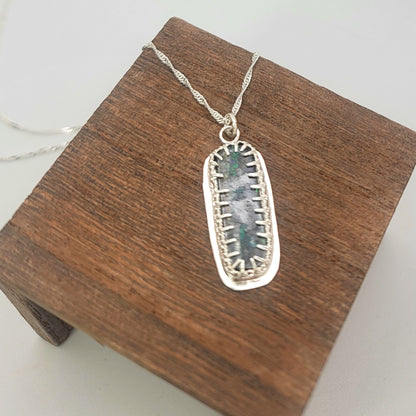 A silver pendant necklace with a textured, rectangular iridescent stone inlay, displayed on a dark brown wooden block against a light gray background.