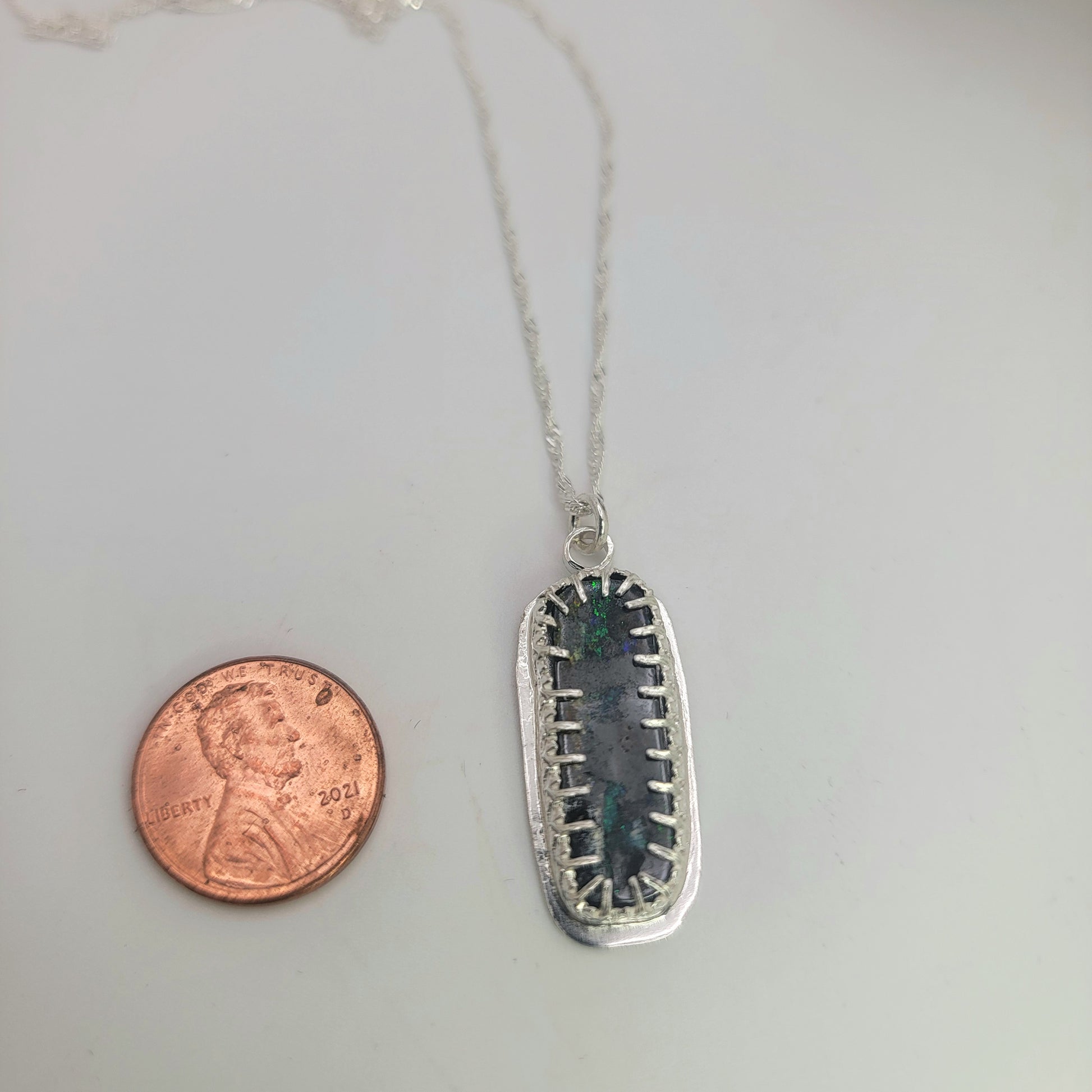 A silver pendant necklace with a rectangular bezel setting holding a dark iridescent stone, shown next to a penny for scale