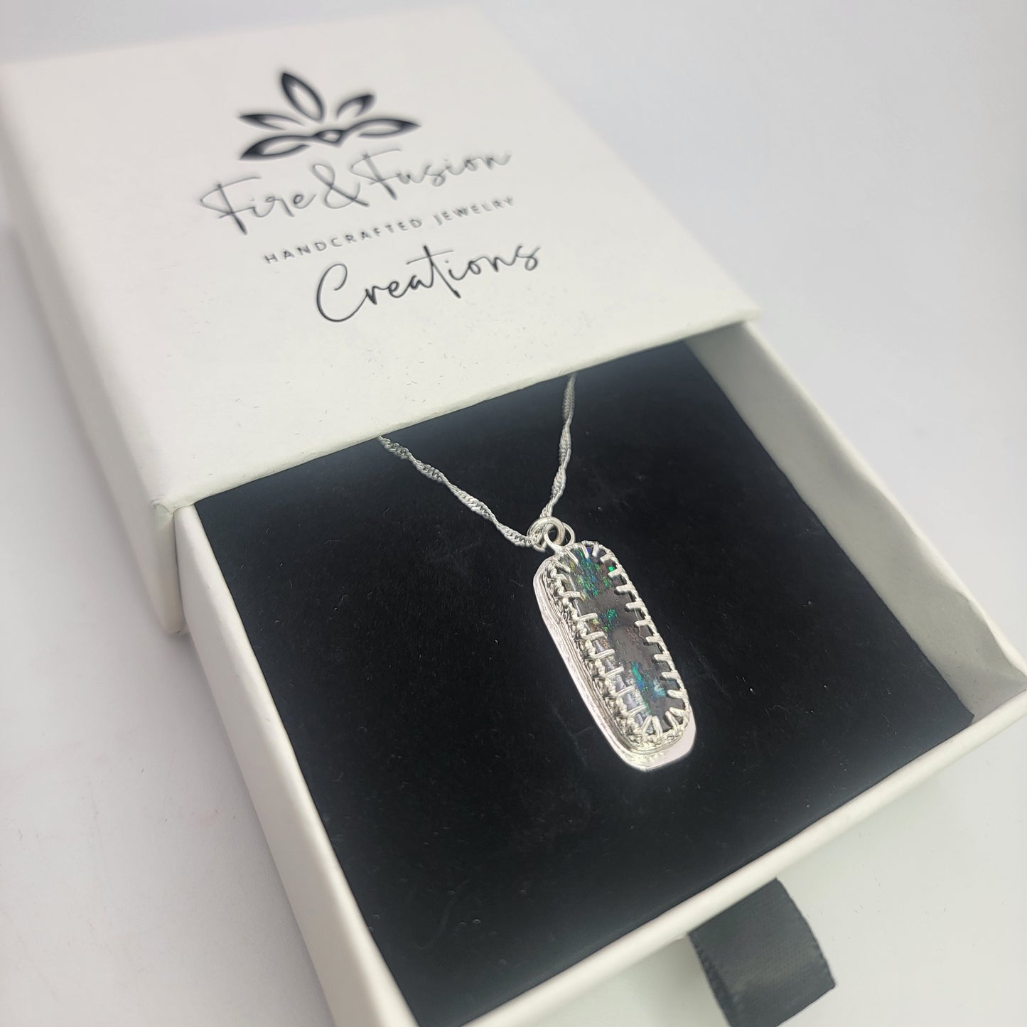 A silver rectangular pendant necklace with a textured border and a central blue-green stone, displayed inside an open white jewelry box with a black velvet insert. The box lid is printed with the brand name 'Fire & Fusion Creations'.
