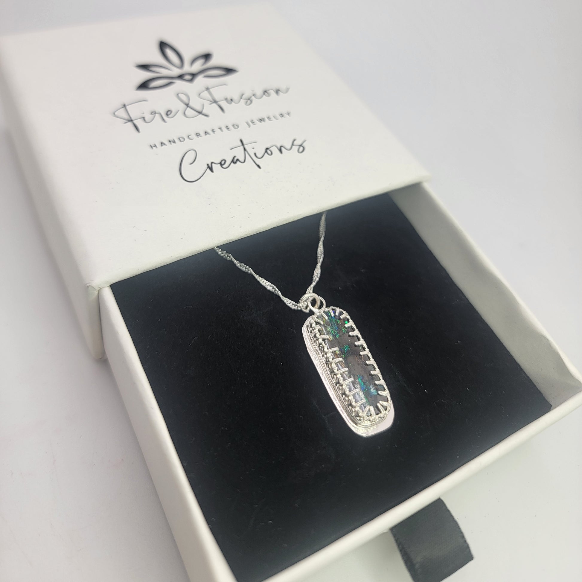 A silver rectangular pendant necklace with a textured border and a central blue-green stone, displayed inside an open white jewelry box with a black velvet insert. The box lid is printed with the brand name 'Fire & Fusion Creations'.