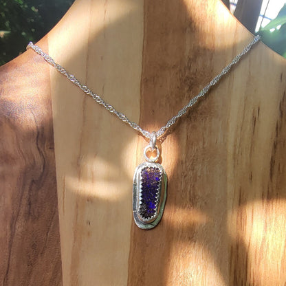 A silver chain necklace with a polished oval pendant featuring a textured purple and blue stone inlay, displayed on a wooden bust with warm sunlight and shadows.