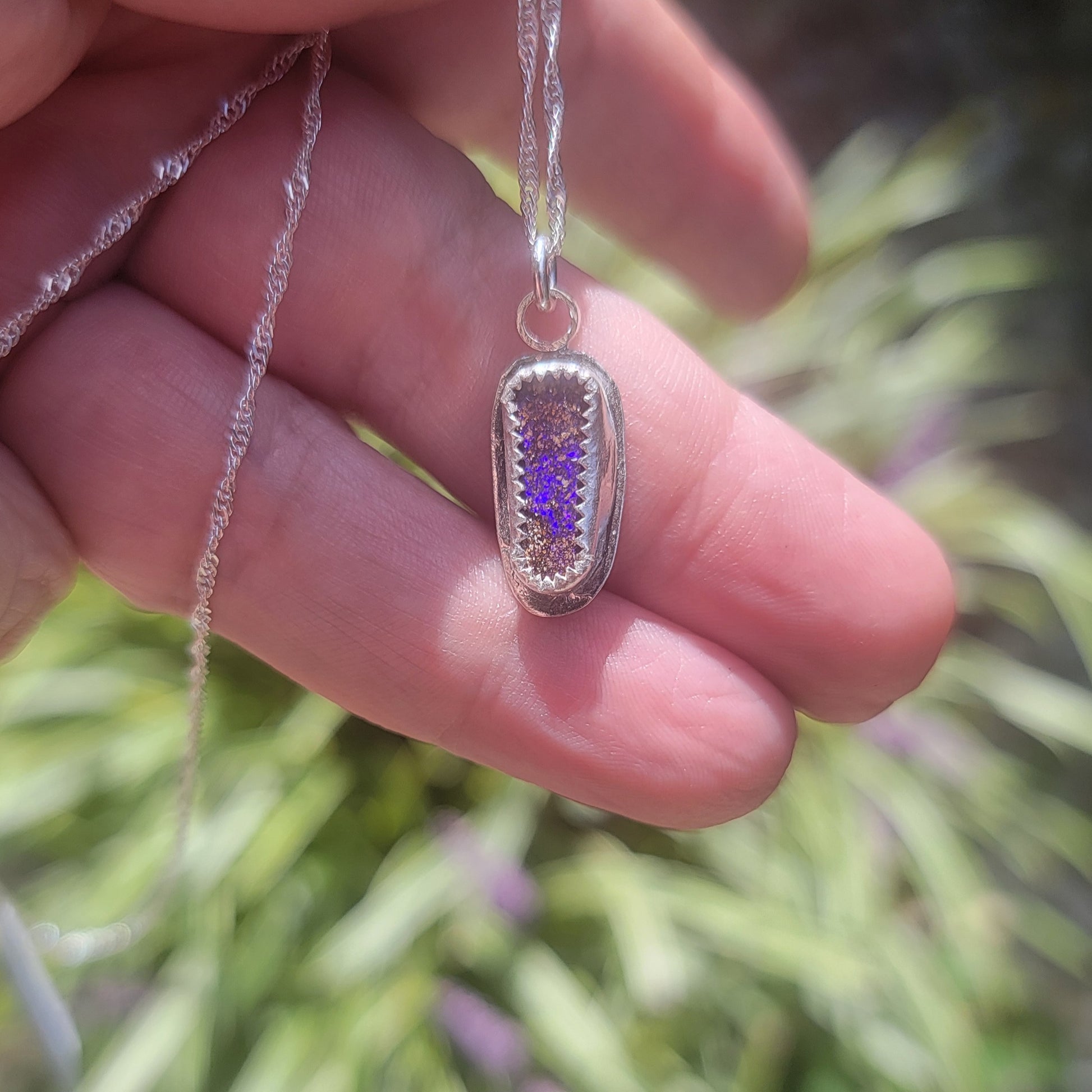 A close-up of a hand holding a silver pendant necklace with a textured, hammered finish and a small purple crystal or enamel center, set against a blurred green outdoor background.