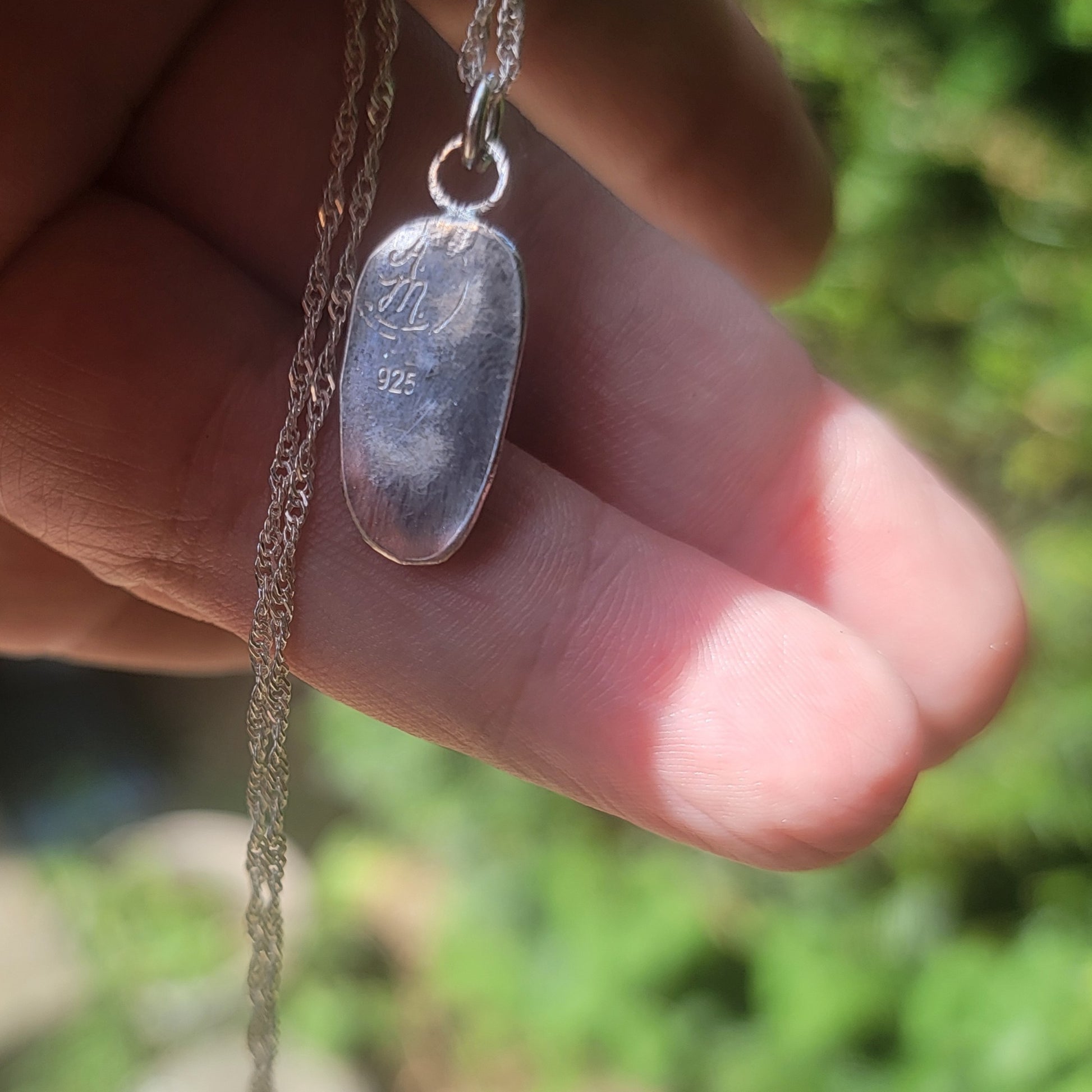 A sterling silver pendant necklace engraved with 'In J. 925' and held in a hand against a blurred green outdoor background.