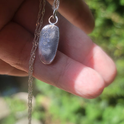 A sterling silver pendant necklace engraved with 'In J. 925' and held in a hand against a blurred green outdoor background.