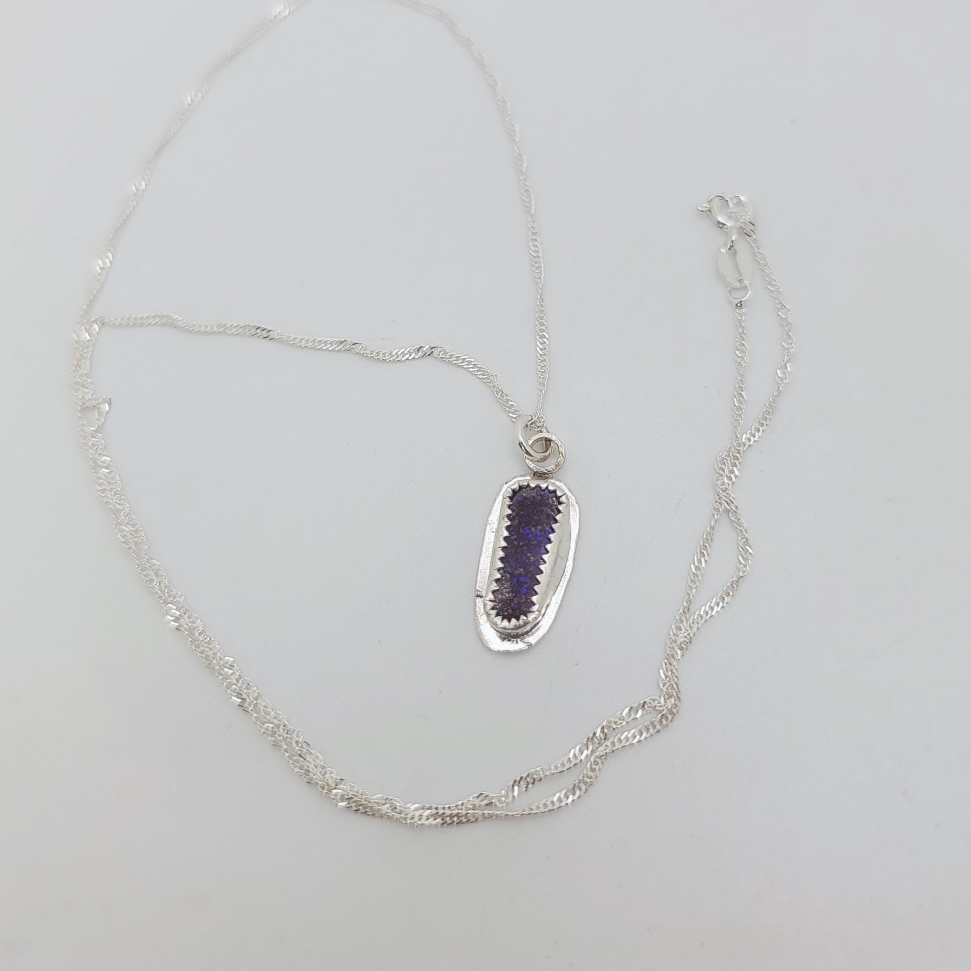 A silver chain necklace featuring an elongated oval pendant with a textured silver border and a central purple amethyst crystal, displayed on a light gray background.