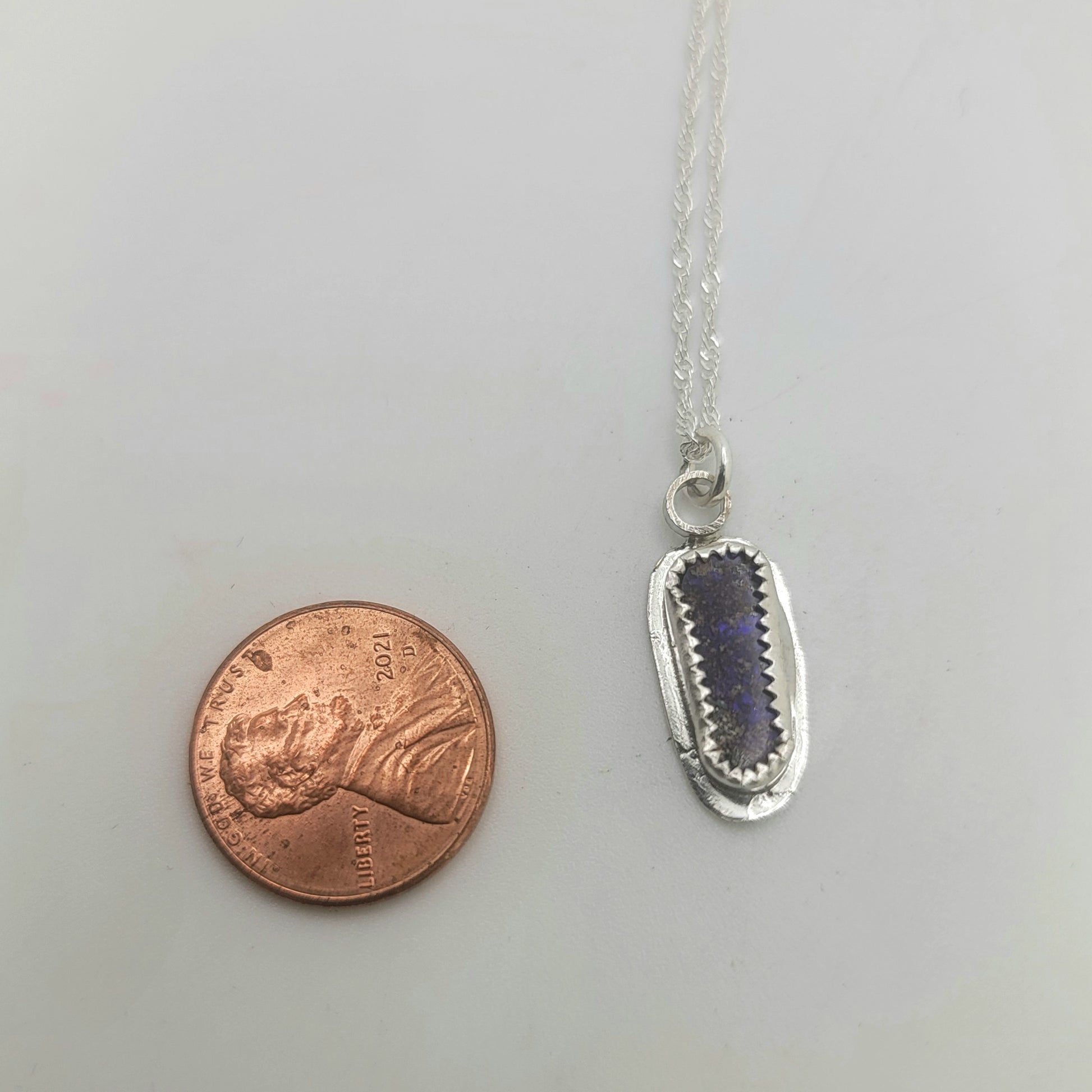 A silver pendant necklace with a dark purple rectangular stone, shown next to a copper penny for scale on a light gray background.