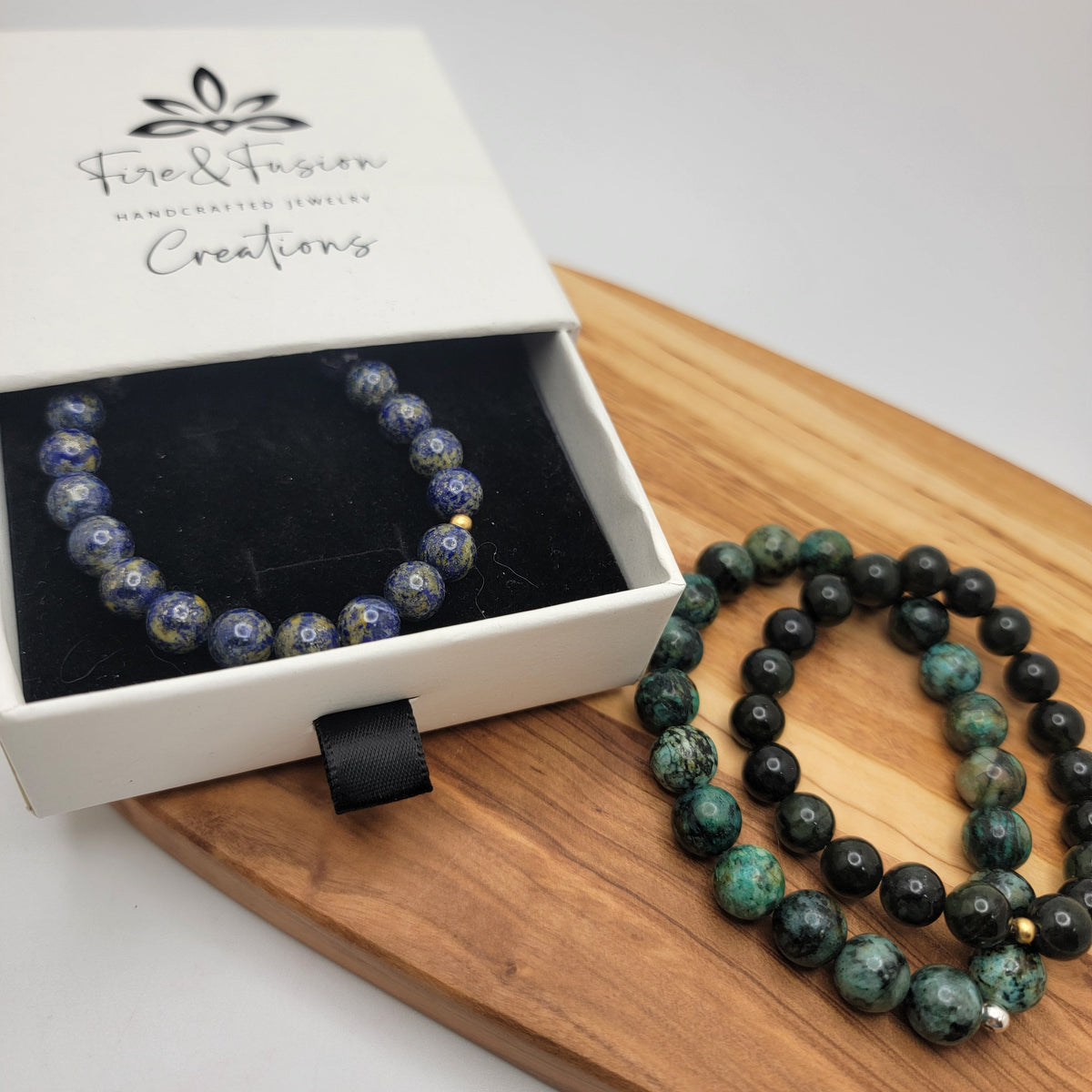 Two beaded bracelets displayed on a wooden board next to a white gift box. One bracelet is blue with gold flecks, and the other is green and black marbled.